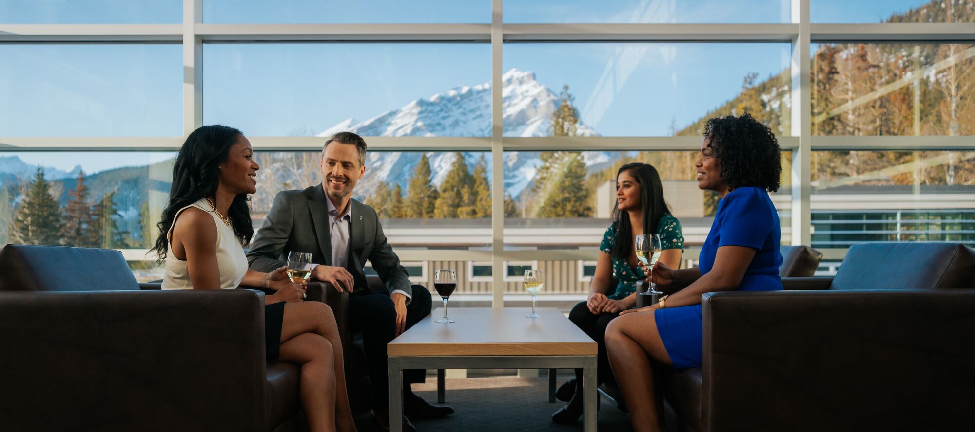 Four people sit in leather chairs at the Banff Centre with mountains in the background while enjoying a glass of wine during an event in Banff National Park.